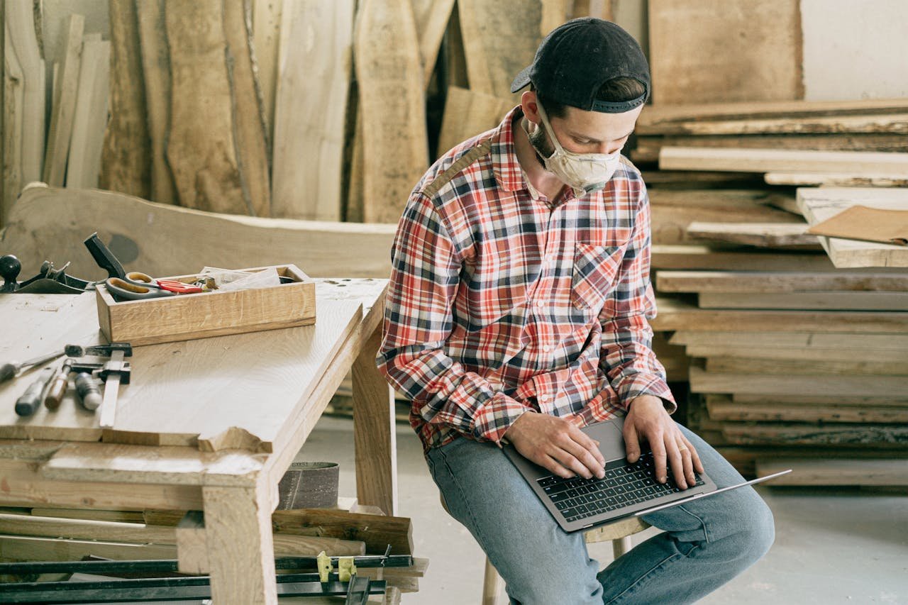 A carpenter in a workshop using a laptop, surrounded by tools and wood.