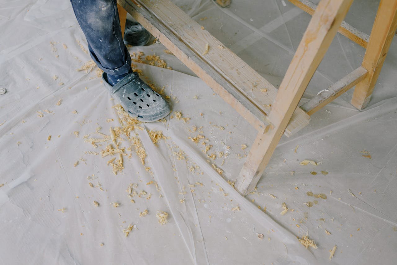 Woodworker in workshop wearing protective footwear surrounded by wood shavings.