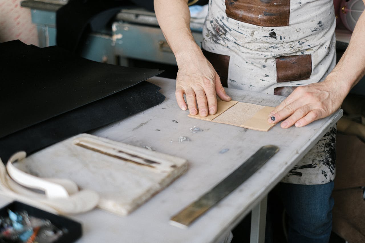Close-up of a craftsman's hands working on leather goods in a workshop setting.